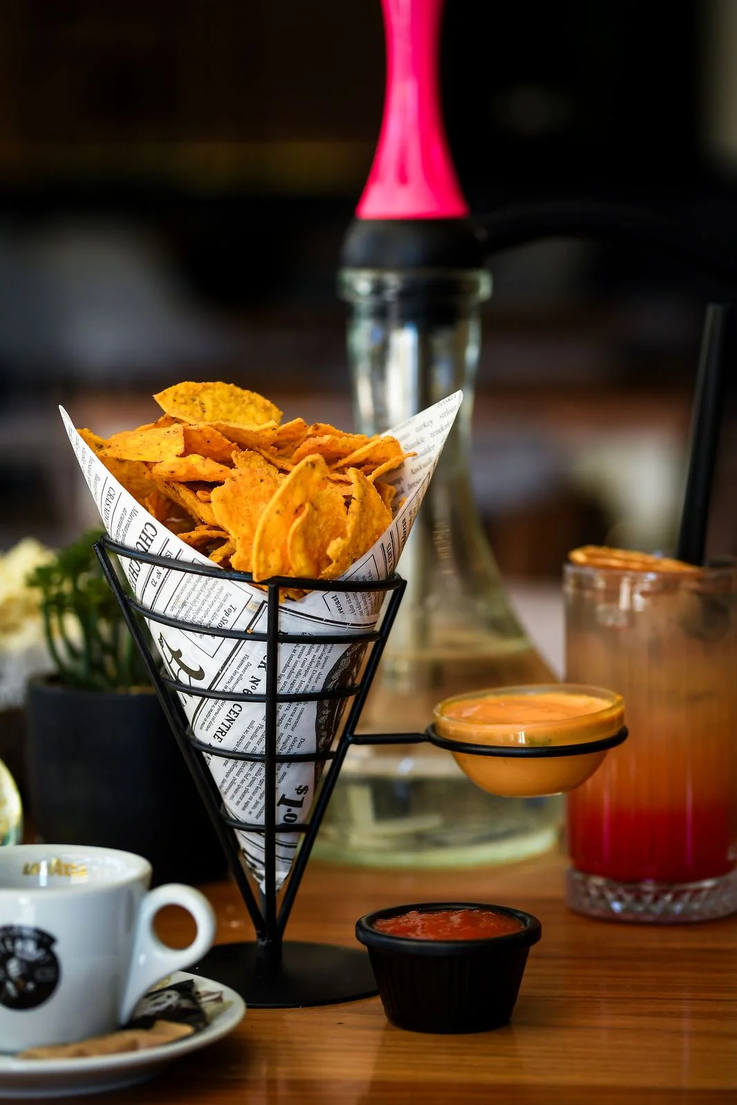 Nachos in a newspaper-print paper cone with two sauces, an espresso and a cocktail, a hookah in the background