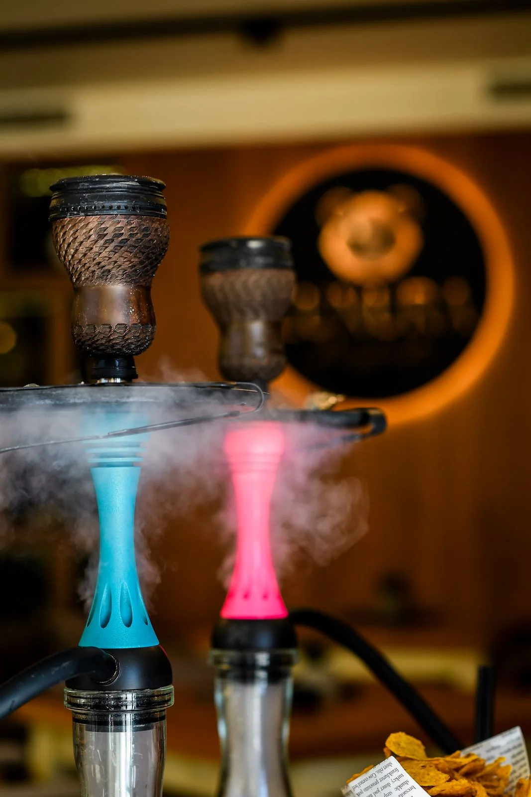 Turquoise and pink hookahs up close, clay bowls on top, the round LUXURA sign blurred in the background