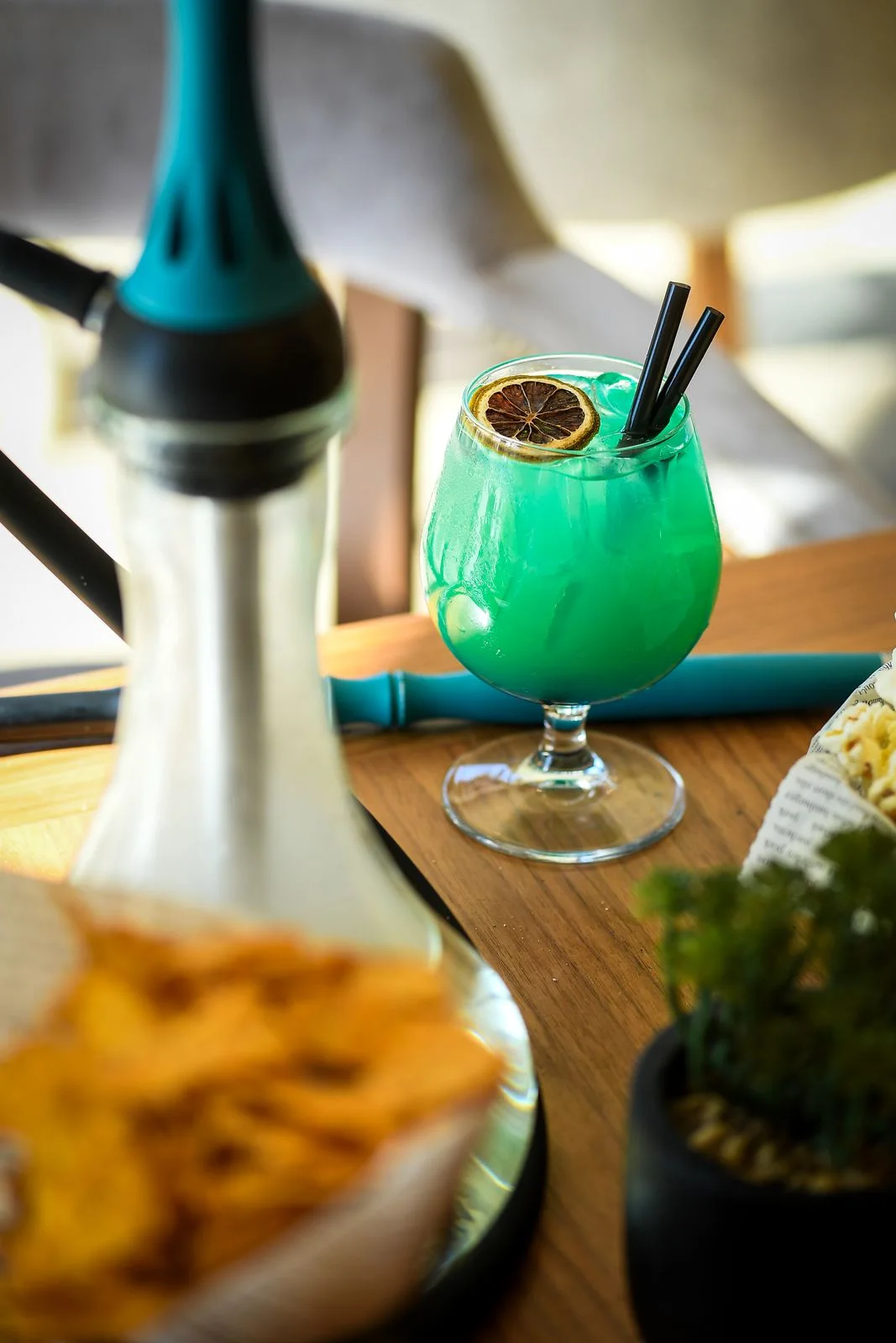 Turquoise cocktail with dried lime in a rounded glass on a wooden table, a hookah blurred in the foreground