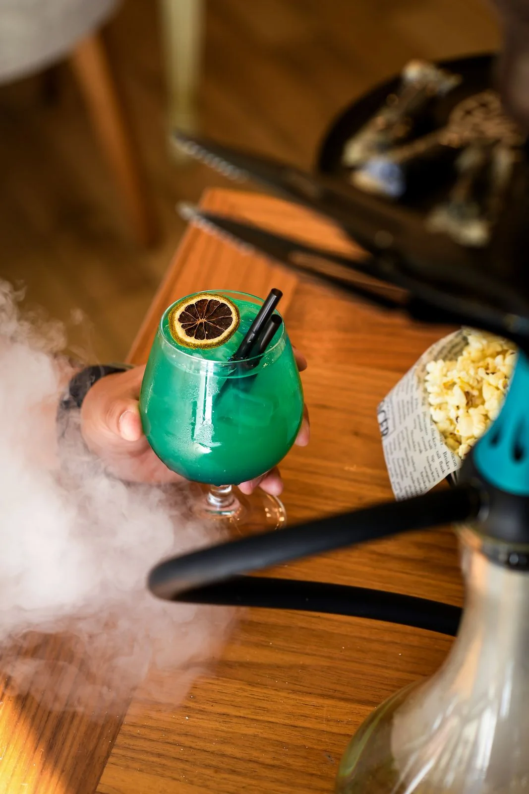 A hand holding a turquoise cocktail with dried lime above a wooden table, popcorn and a hookah to the side