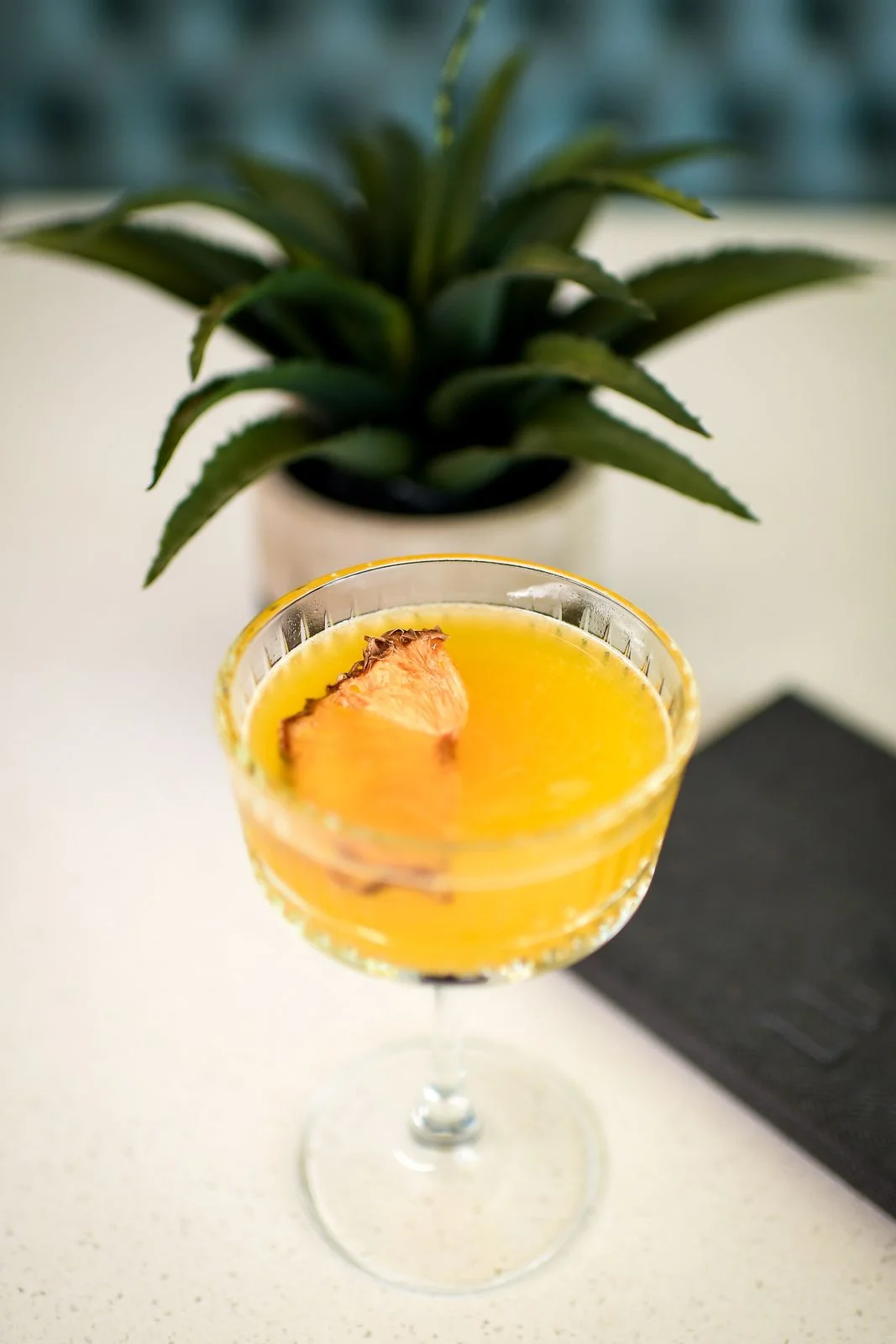 Yellow cocktail in a coupe glass with a slice of dried pineapple, a plant and a black menu on a white counter