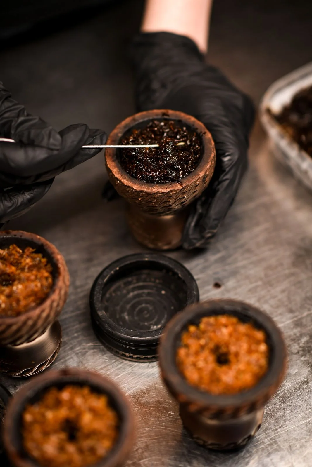 Hands in black gloves spreading dark tobacco in a clay hookah bowl with a needle