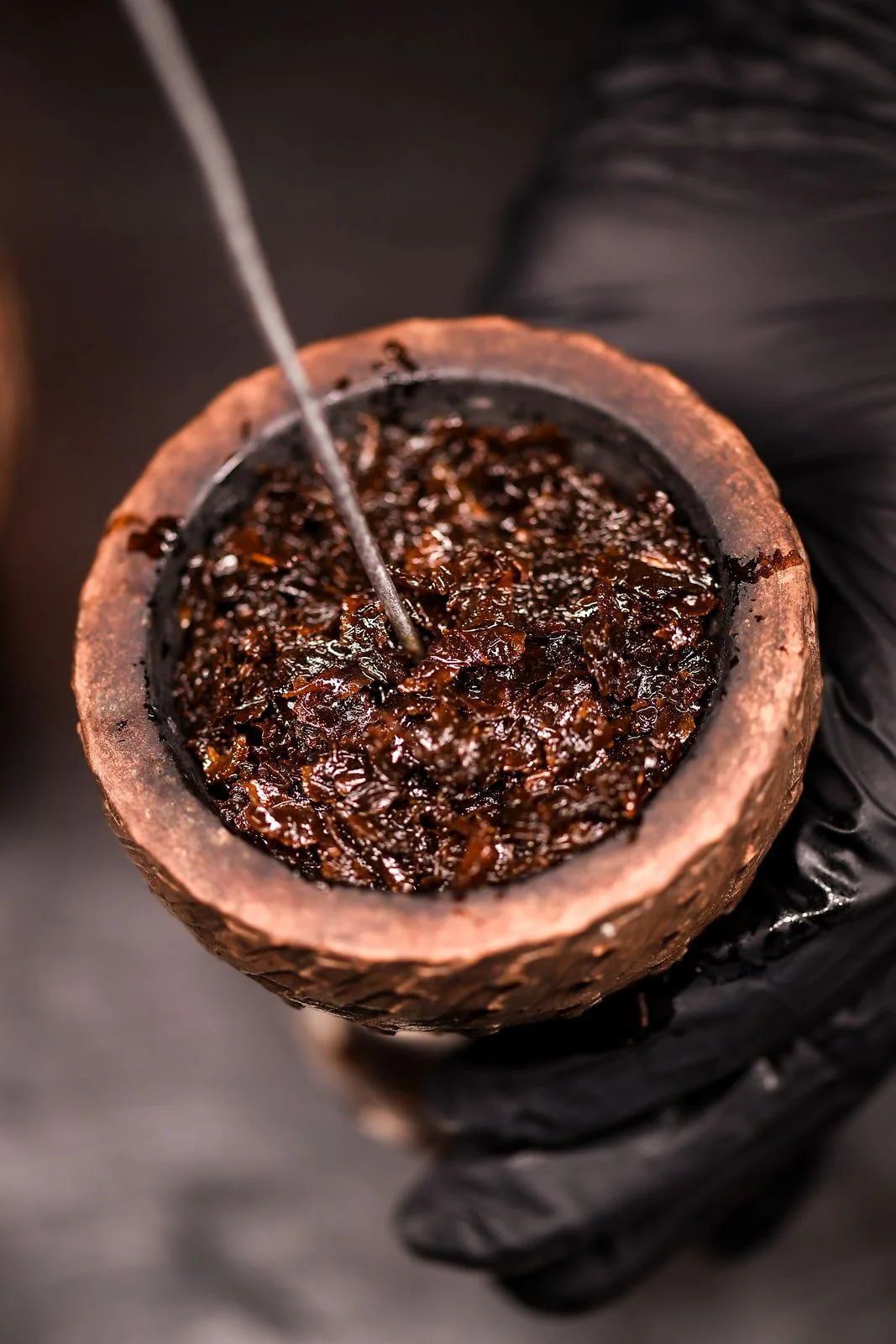 Close-up: a needle loosening dark tobacco in a clay bowl held by a gloved hand