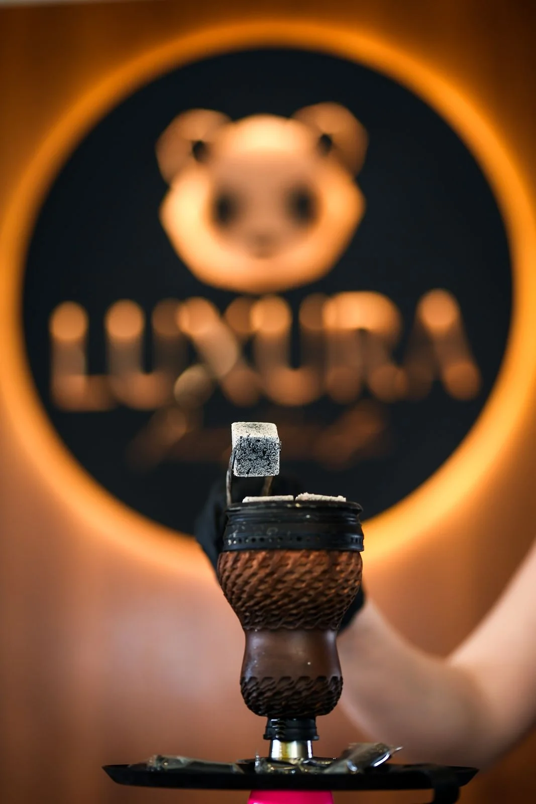 A gloved hand using tongs to place coals onto a clay hookah bowl, the LUXURA logo blurred behind