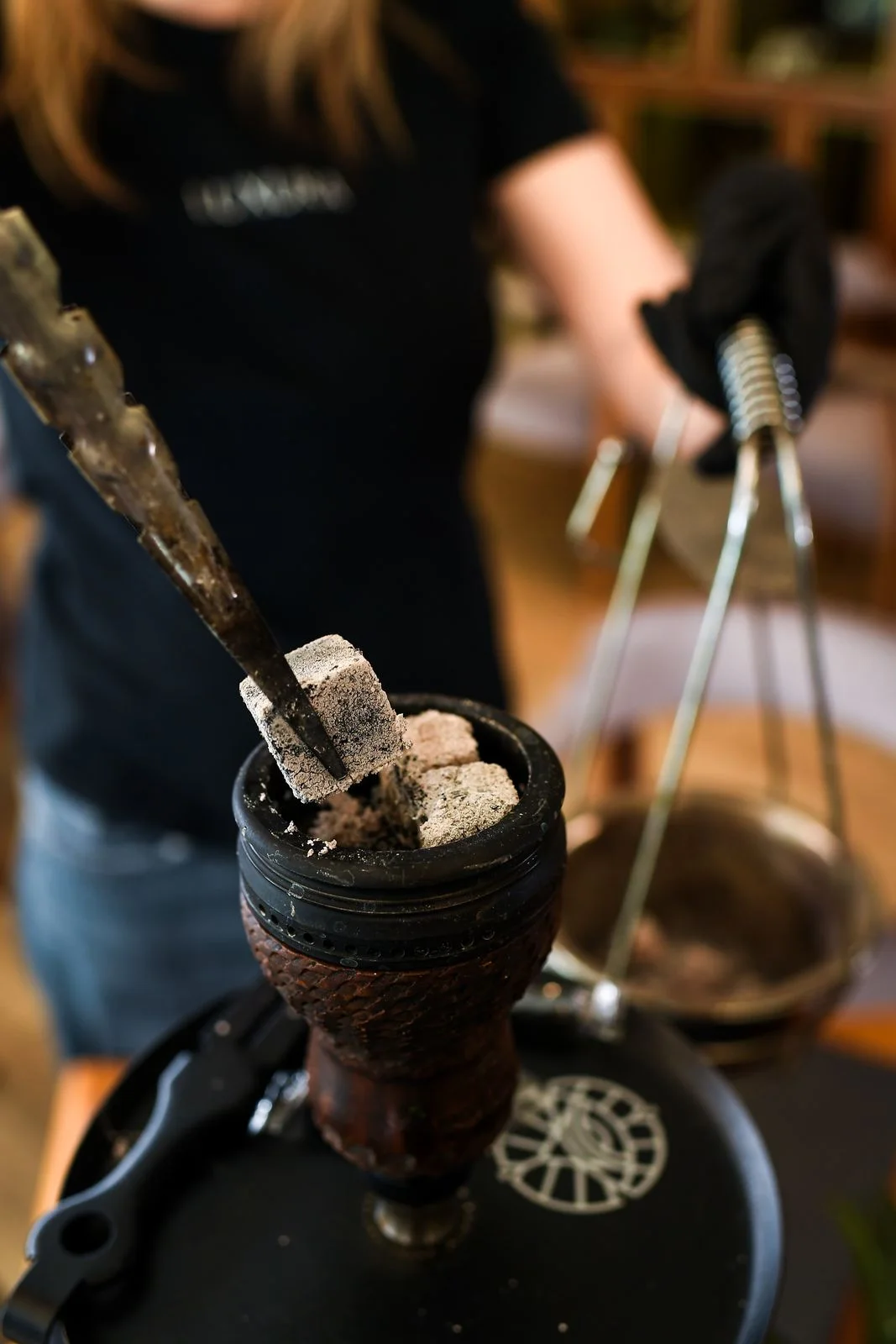 A staff member in a black LUXURA T-shirt arranging coals on a hookah bowl with tongs