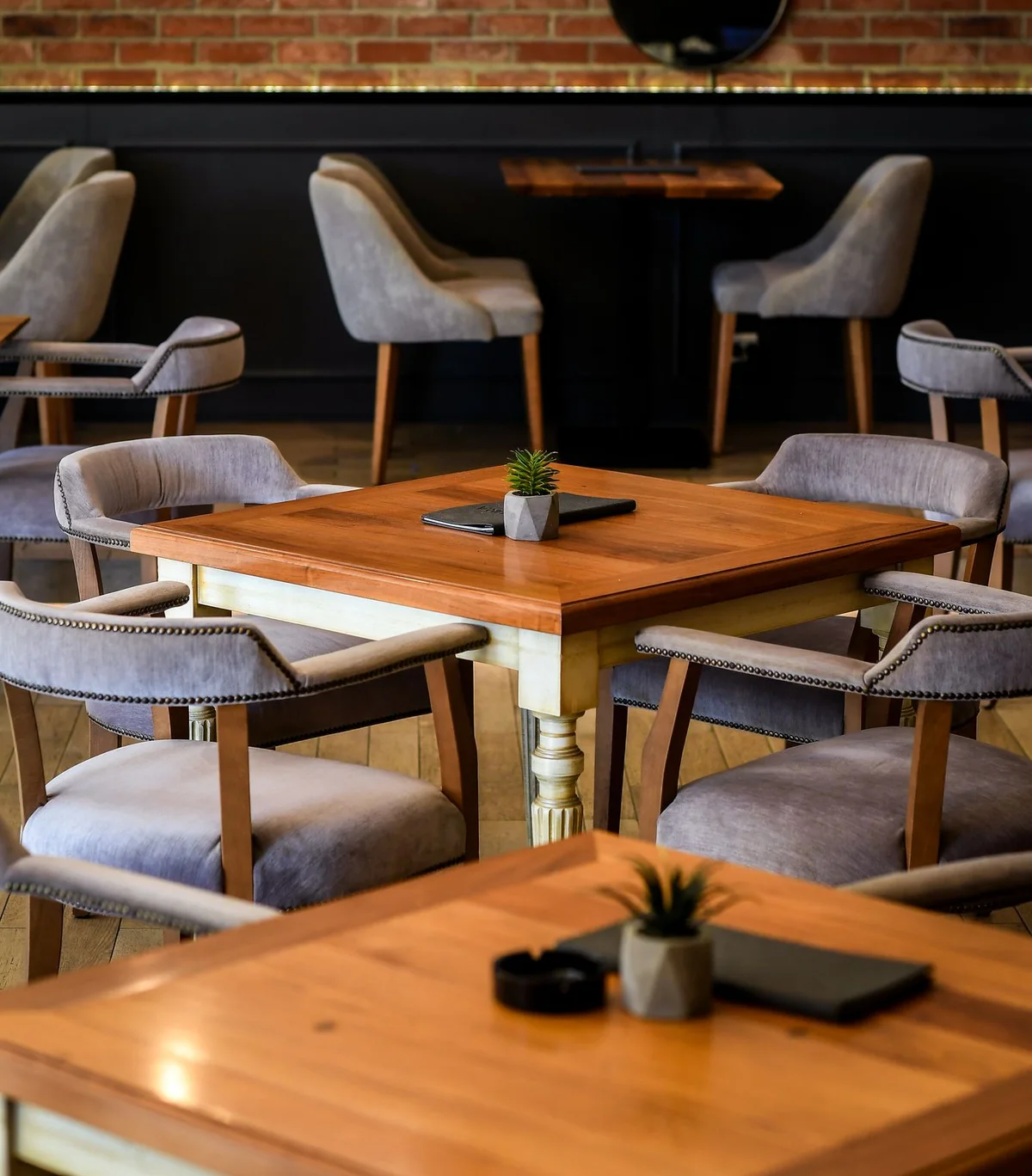 Square wooden table with a succulent and gray velvet armchairs with decorative studs, brick behind