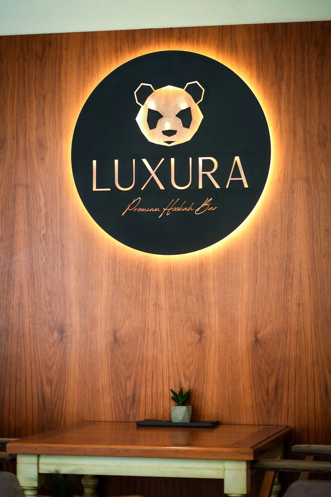 Illuminated round LUXURA Premium Hookah Bar sign with the panda on a walnut wall, a wooden table below