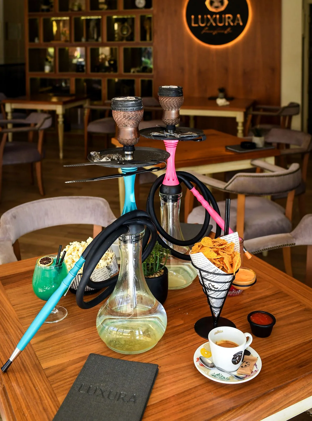 Two hookahs, turquoise and pink, with a cocktail, popcorn, nachos and an espresso; the illuminated LUXURA sign behind
