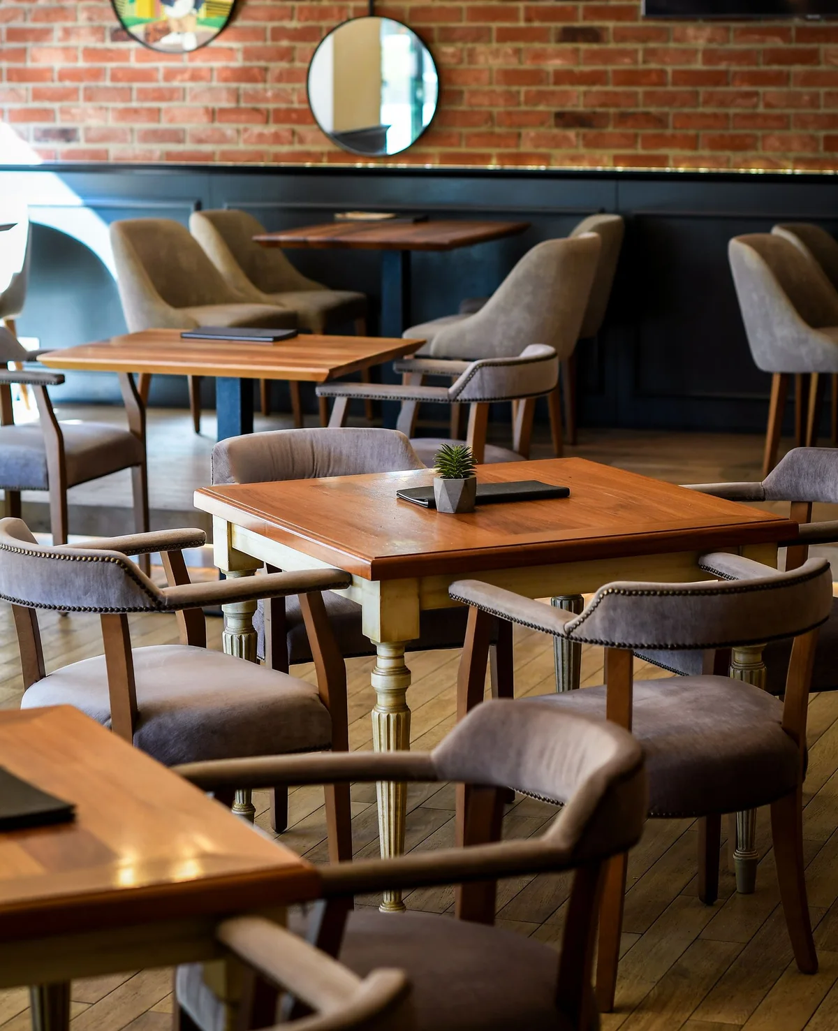 Inside the LUXURA café in A blok: wooden tables, gray armchairs and a round mirror on a brick wall