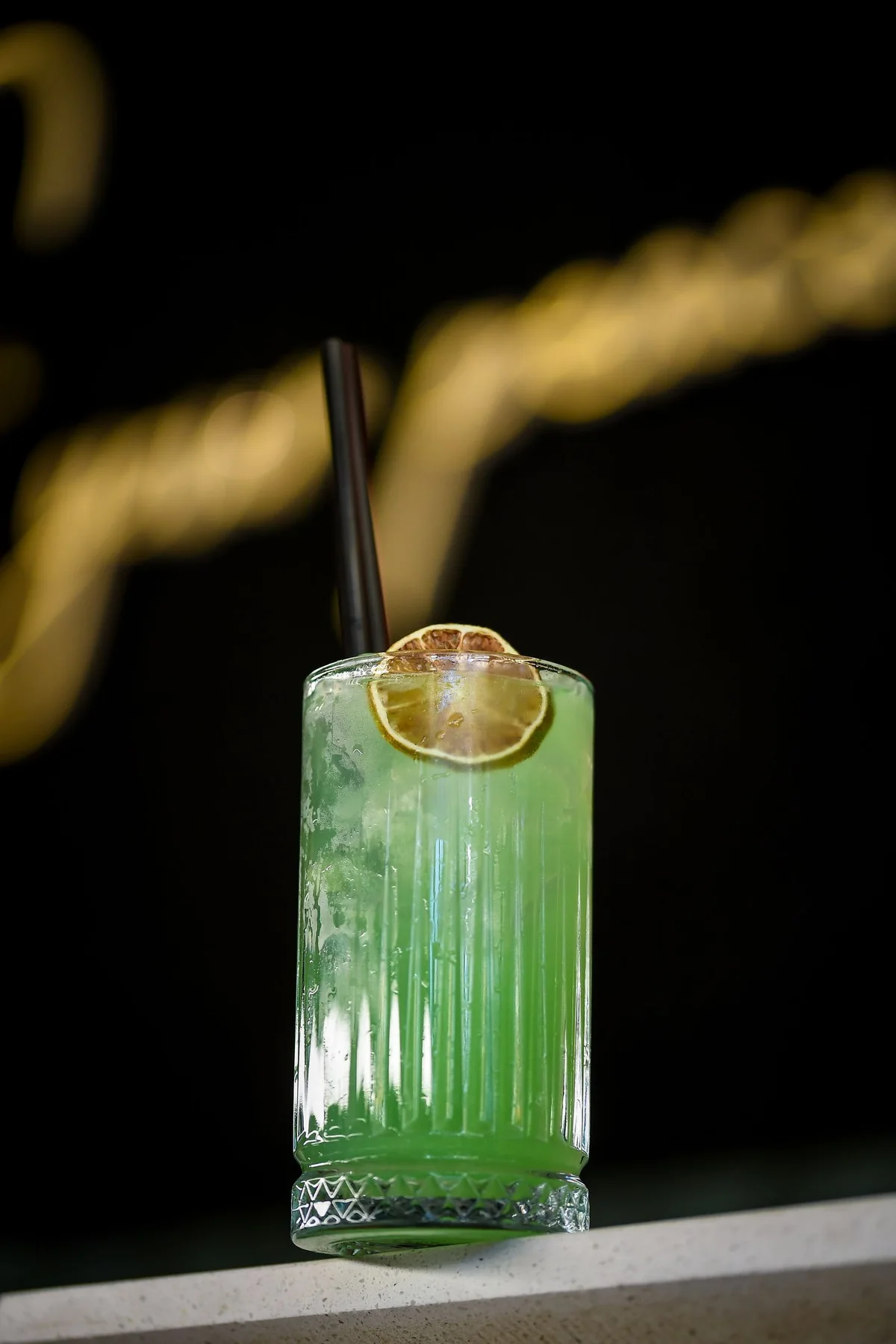Green cocktail in a ribbed glass with dried lime and a black straw, a gold neon sign blurred behind