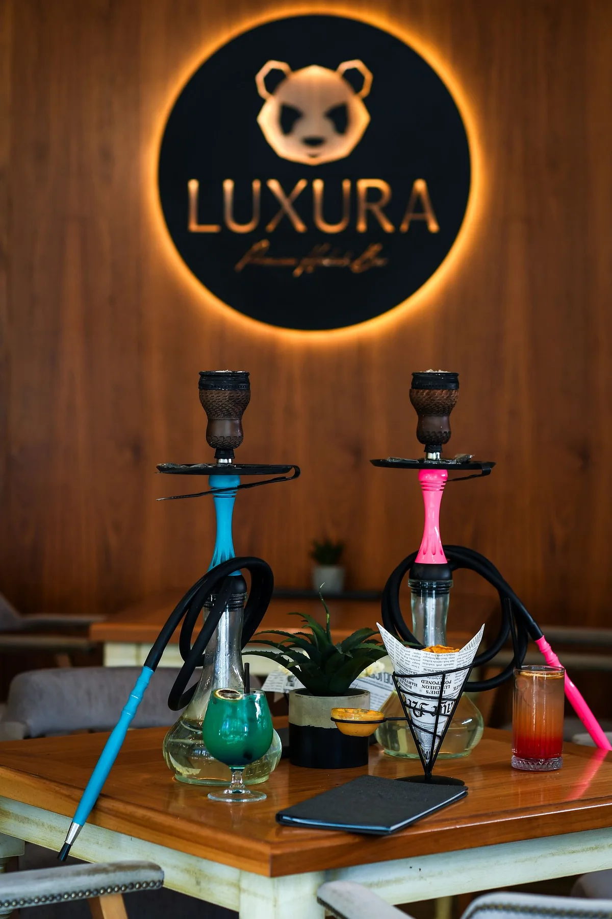 LUXURA hookah bar: turquoise and pink hookahs, two cocktails and nachos on a table in front of the illuminated logo