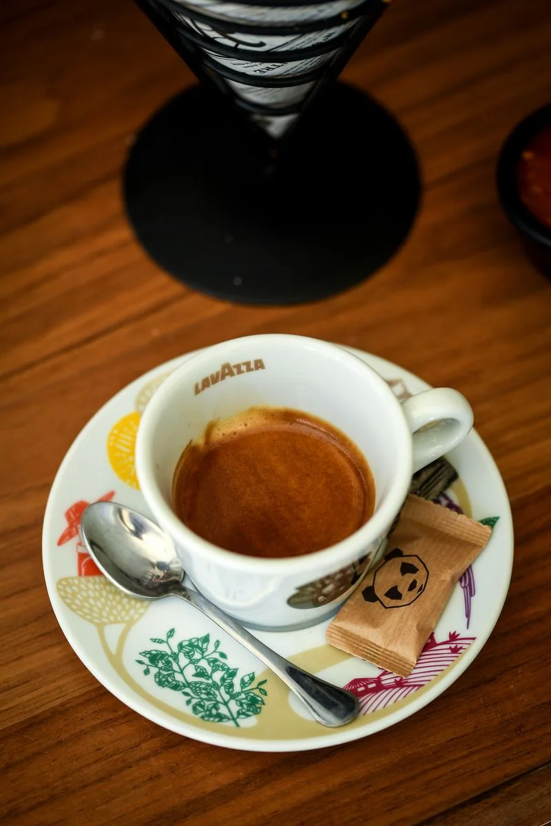 Espresso in a white cup on a patterned saucer, a teaspoon and a sugar sachet with the LUXURA panda