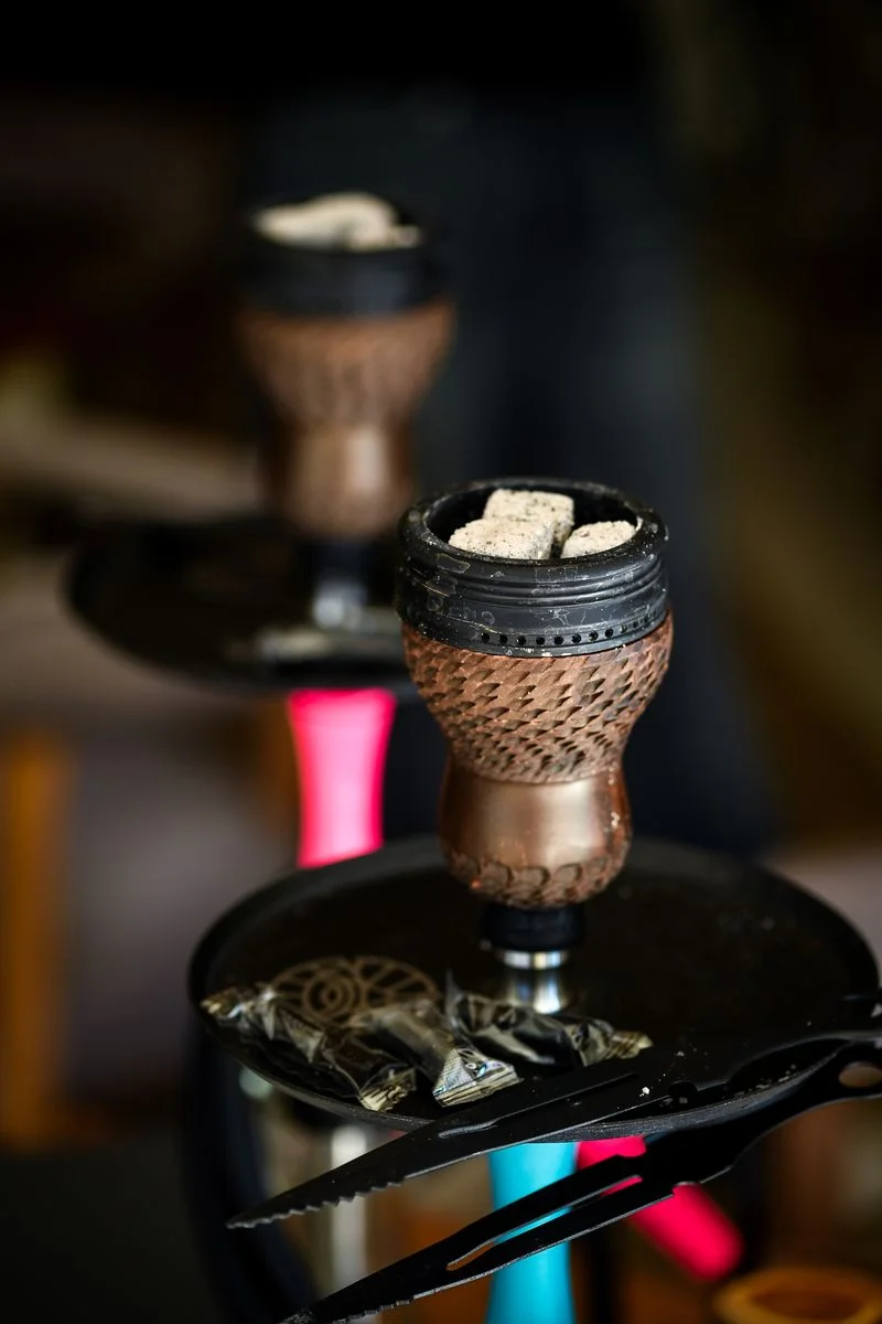 Hookah at LUXURA: a clay bowl with charcoal cubes on a black tray, another hookah blurred behind