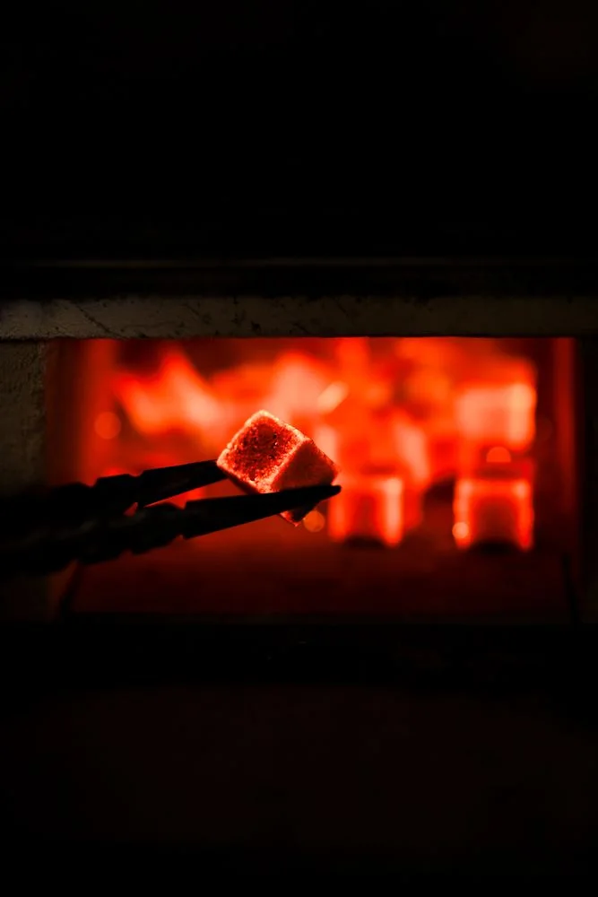 A glowing hookah charcoal cube in tongs, a coal heater full of hot coals in the background