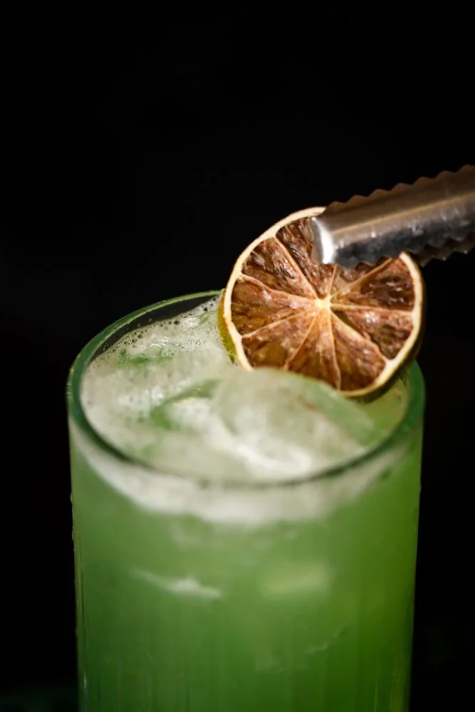 A dried lime wheel in tongs on the rim of a glass with a green cocktail, close-up